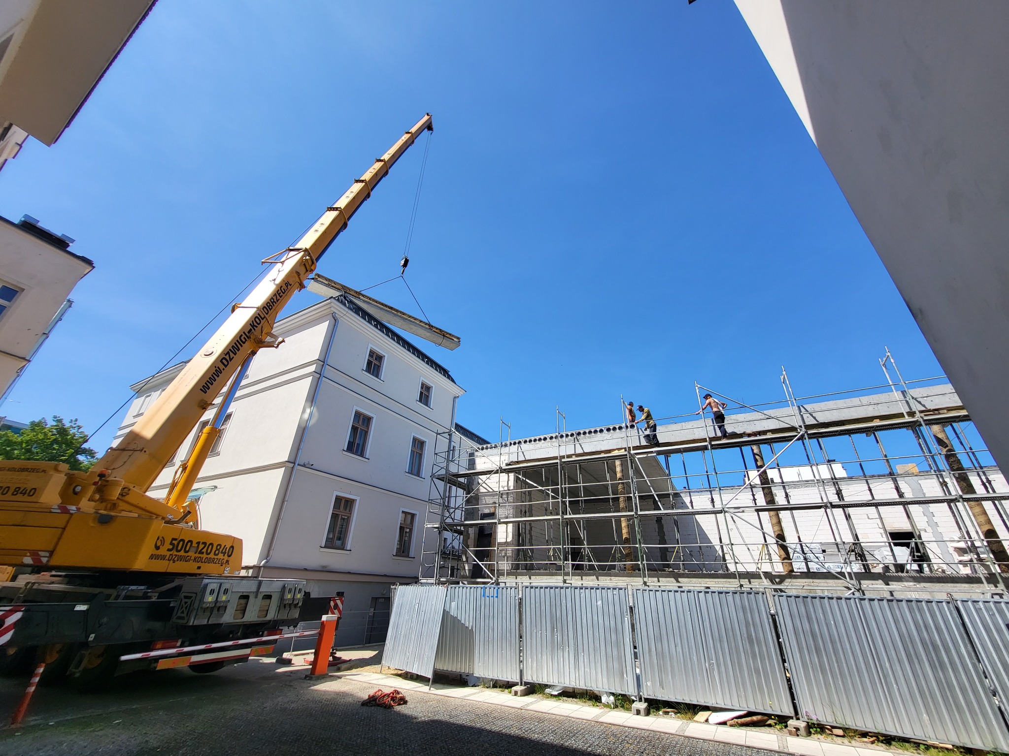 Montaż stropu budynku z użyciem dźwigu, widoczne rusztowania i pracownicy. Ujęcie z dołu, błękitne niebo w tle. Dźwig firmy Dźwigi-Kołobrzeg.