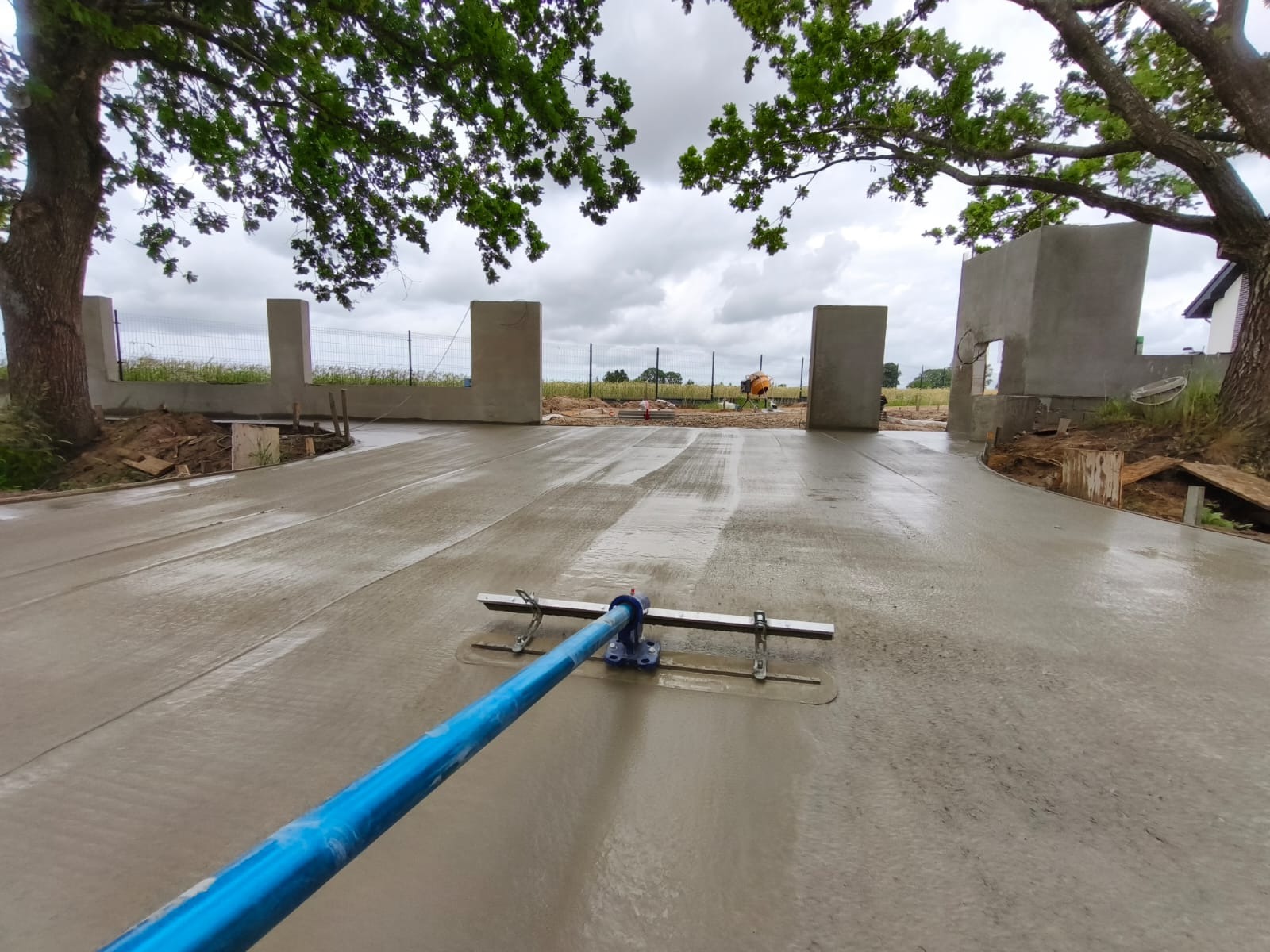 Świeżo wylana, mokra wylewka betonowa na posesji, wyrównywana narzędziem z długim, niebieskim uchwytem. W tle słupki ogrodzenia i fragmenty konstrukcji budynku.