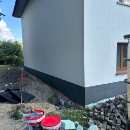 OLBUD OLEH BEZHUK - Fragment elewacji budynku z wykończoną dolną częścią w kolorze grafitowym. Widoczne narzędzia i materiały budowlane, w tym puszki z tynkiem i styropian.