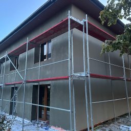 OLBUD OLEH BEZHUK - Dom w trakcie tynkowania elewacji, widoczne rusztowania i świeżo nałożony tynk w odcieniach szarości. Częściowo ocieplony budynek z zamontowanymi oknami i drzwiami.