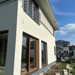 OLBUD OLEH BEZHUK - Jasna elewacja domu jednorodzinnego z drewnianą stolarką okienną i metalową balustradą. Widoczni pracownicy przy elewacji. Ujęcie z dołu, błękitne niebo.