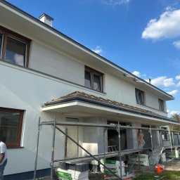 OLBUD OLEH BEZHUK - Jasny, dwukondygnacyjny dom z rusztowaniem i farbami, w trakcie prac elewacyjnych. Widoczne puszki z farbą i materiały izolacyjne. Błękitne niebo w tle.