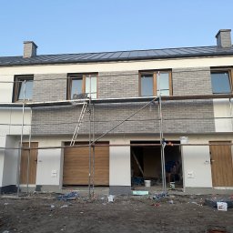 OLBUD OLEH BEZHUK - Elewacja szeregowca w trakcie wykończenia: szare płytki imitujące cegłę na fragmencie ściany, rusztowania, widoczne puszki z farbą. Prace wykończeniowe w toku.