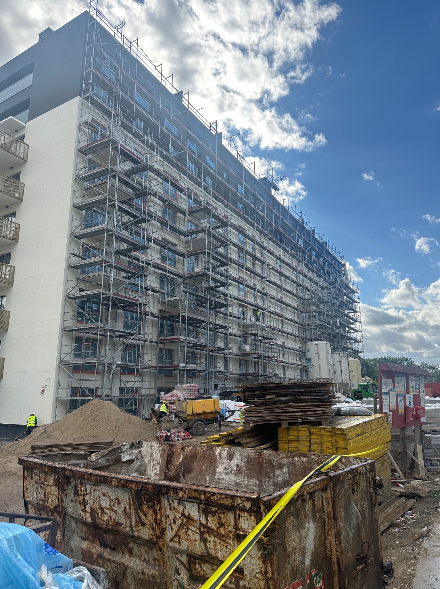 Wielopiętrowy budynek w trakcie prac elewacyjnych, otoczony rusztowaniami. Widoczne materiały budowlane i pracownicy w odblaskowych kamizelkach. Błękitne niebo z chmurami w tle.