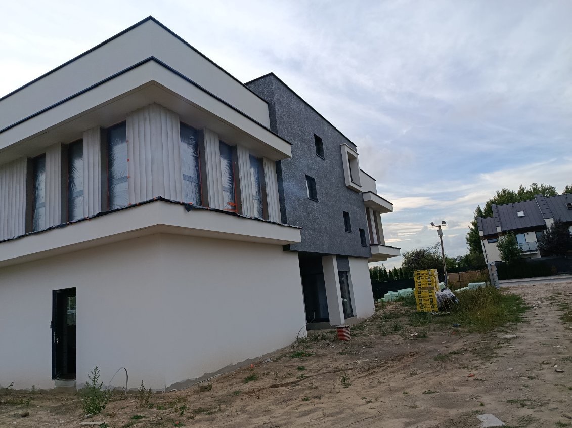 Dwukondygnacyjny dom z szarą elewacją i białymi elementami architektonicznymi. Okna zabezpieczone folią. Widoczne materiały budowlane. Ujęcie z dołu, pochmurne niebo.