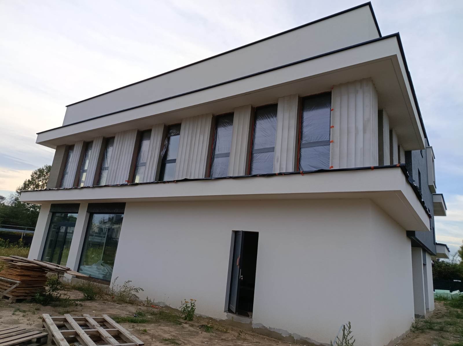 Nowoczesny dom z elewacją w trakcie prac: białe ściany, okna zabezpieczone folią, widoczne elementy dekoracyjne. Palety na ziemi sugerują trwające prace wykończeniowe.