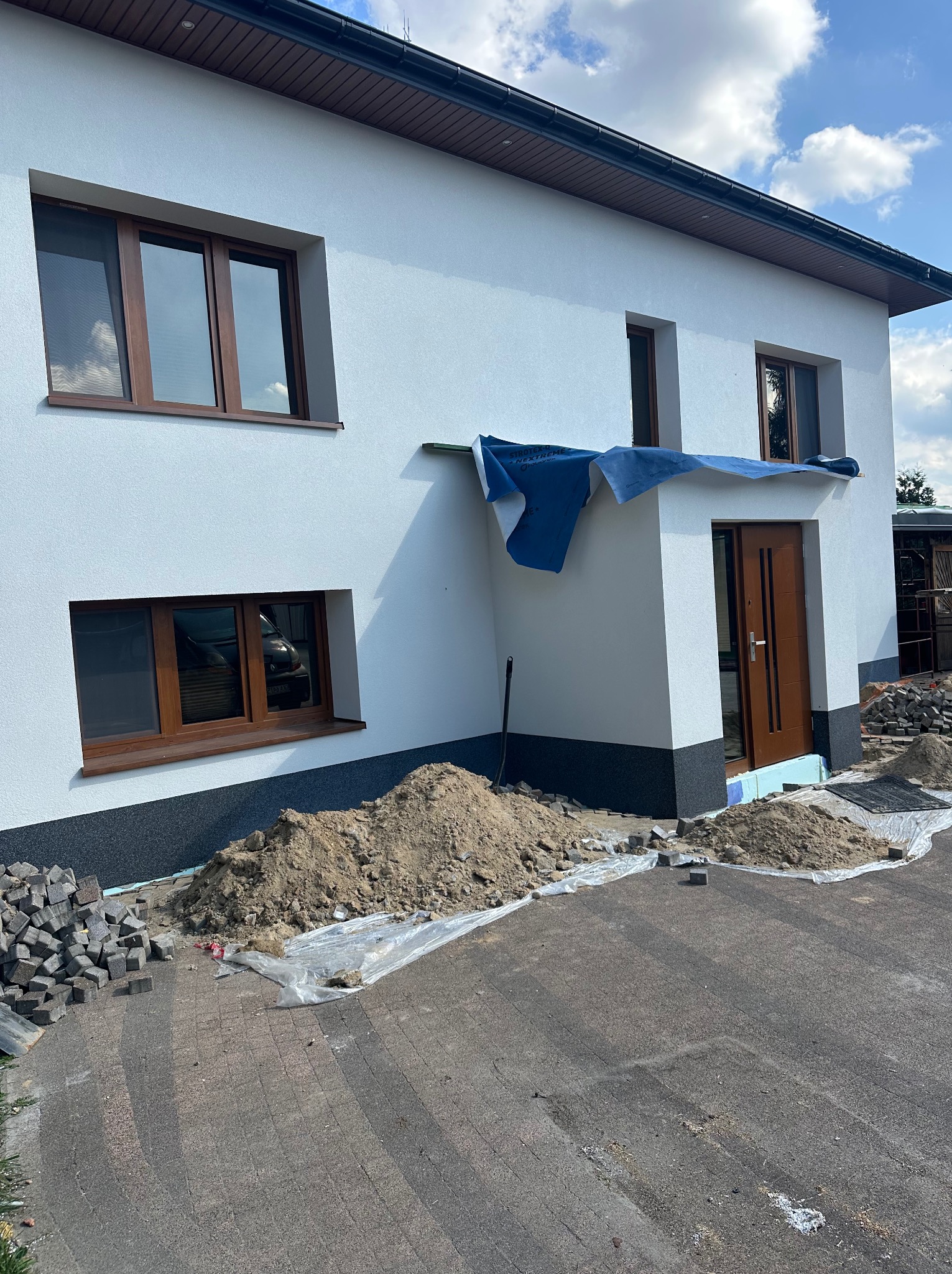 Biały dom z brązowymi oknami w trakcie prac wykończeniowych elewacji i brukowania podjazdu. Widoczne materiały budowlane i zabezpieczenia.