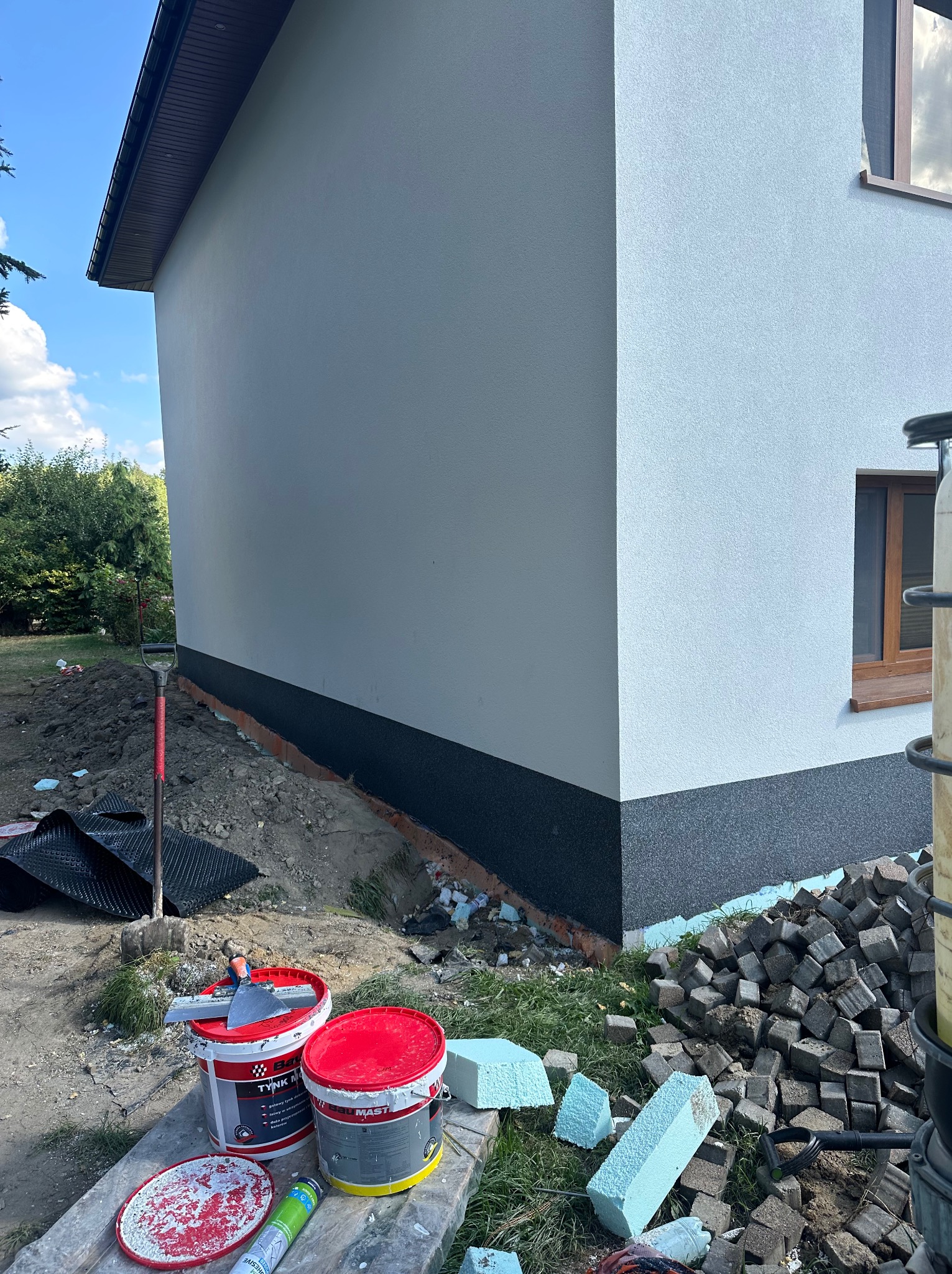 Fragment elewacji budynku z wykończoną dolną częścią w kolorze grafitowym. Widoczne narzędzia i materiały budowlane, w tym puszki z tynkiem i styropian.