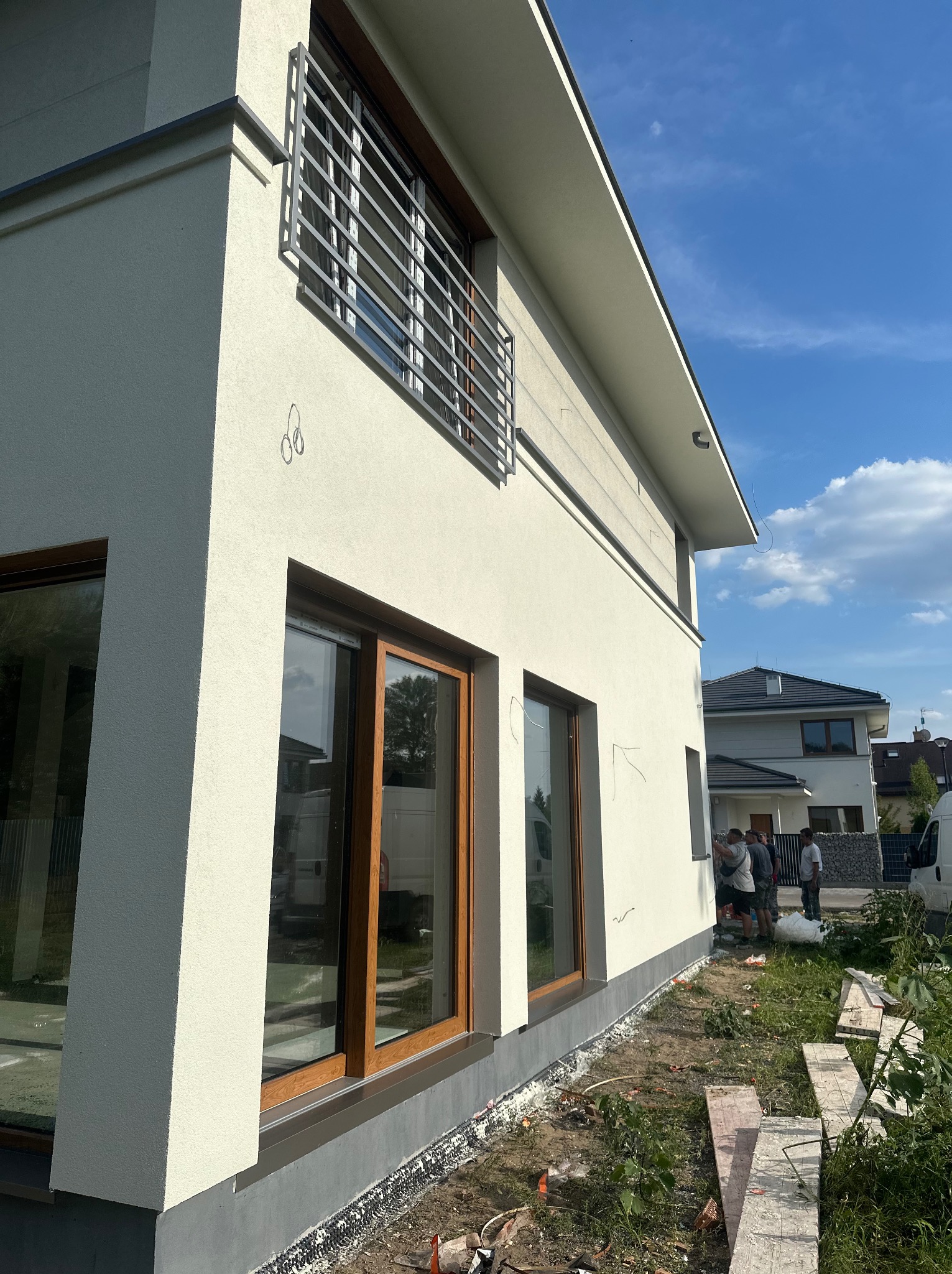 Jasna elewacja domu jednorodzinnego z drewnianą stolarką okienną i metalową balustradą. Widoczni pracownicy przy elewacji. Ujęcie z dołu, błękitne niebo.