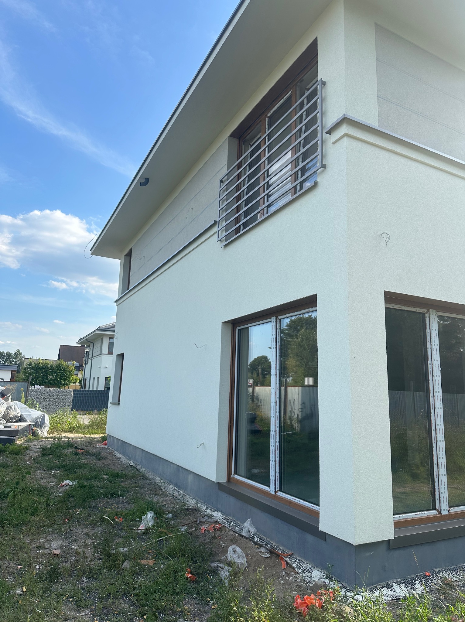 Nowoczesny dom z jasną elewacją i metalową balustradą balkonową. Widoczne duże okna z brązowymi ramami i fragment ogrodu. Minimalistyczny design.