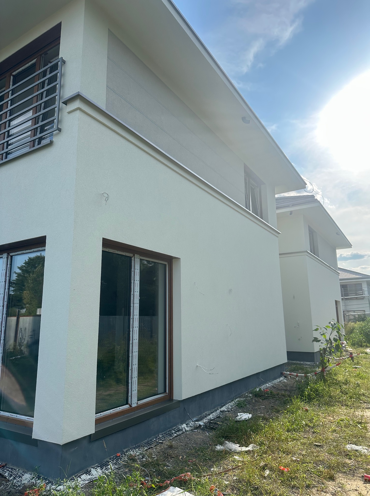 Jasna elewacja dwukondygnacyjnego domu z oknami w brązowych ramach, widok z dołu, fragment trawnika. Elementy wykończeniowe w kolorze szarym.