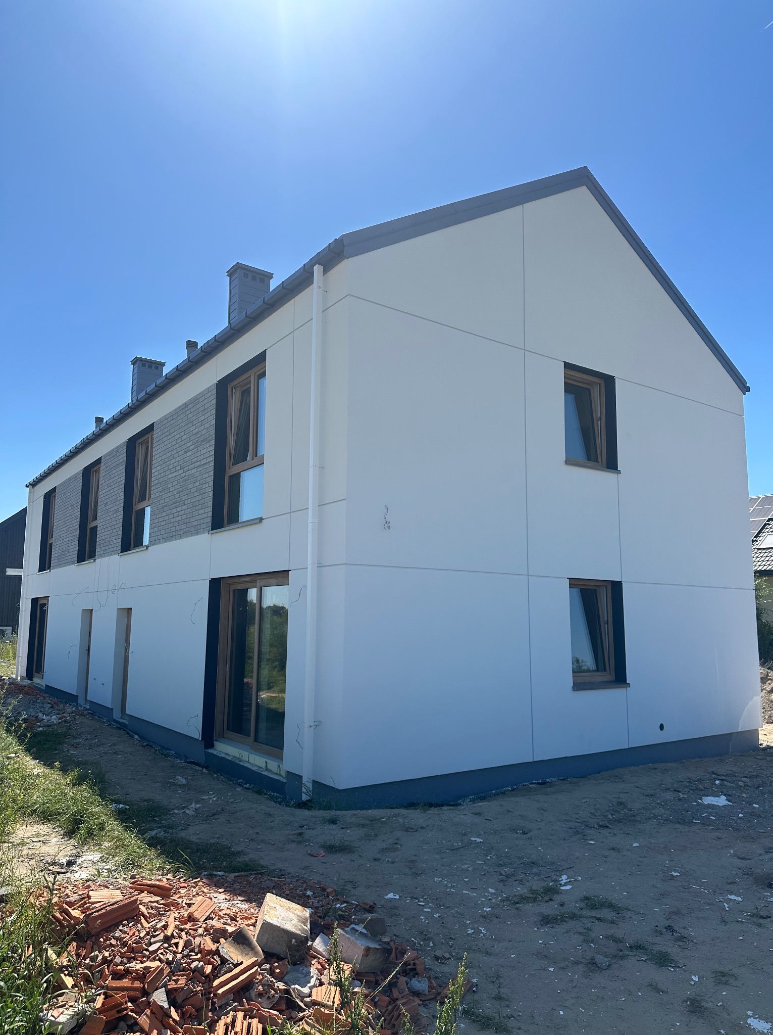 Nowoczesny dom z białą elewacją panelową i szarą cegłą, okna z ciemnymi ramami. Widoczne gruzy i niezagospodarowany teren wokół budynku. Słoneczny dzień.