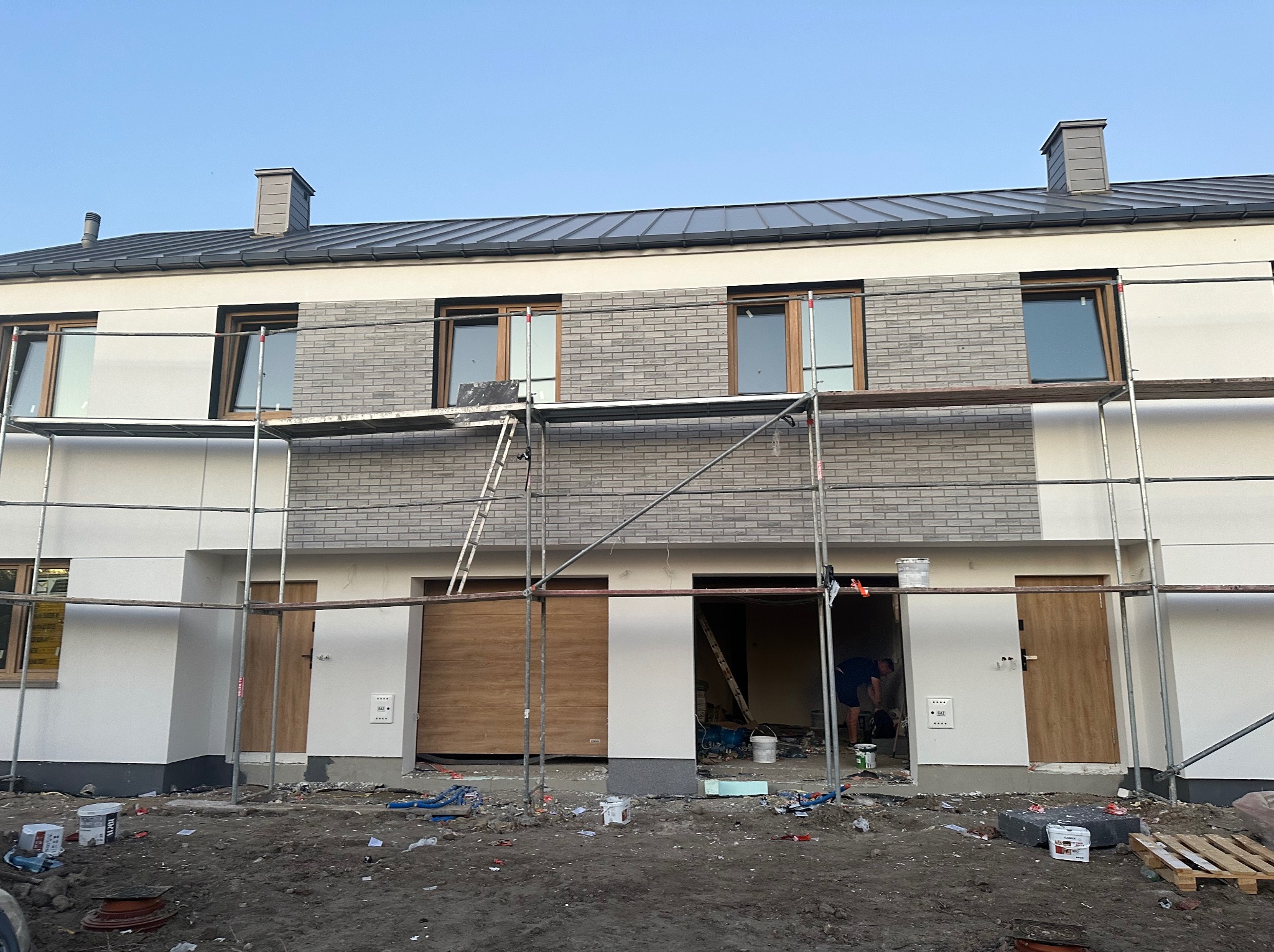 Elewacja szeregowca w trakcie wykończenia: szare płytki imitujące cegłę na fragmencie ściany, rusztowania, widoczne puszki z farbą. Prace wykończeniowe w toku.