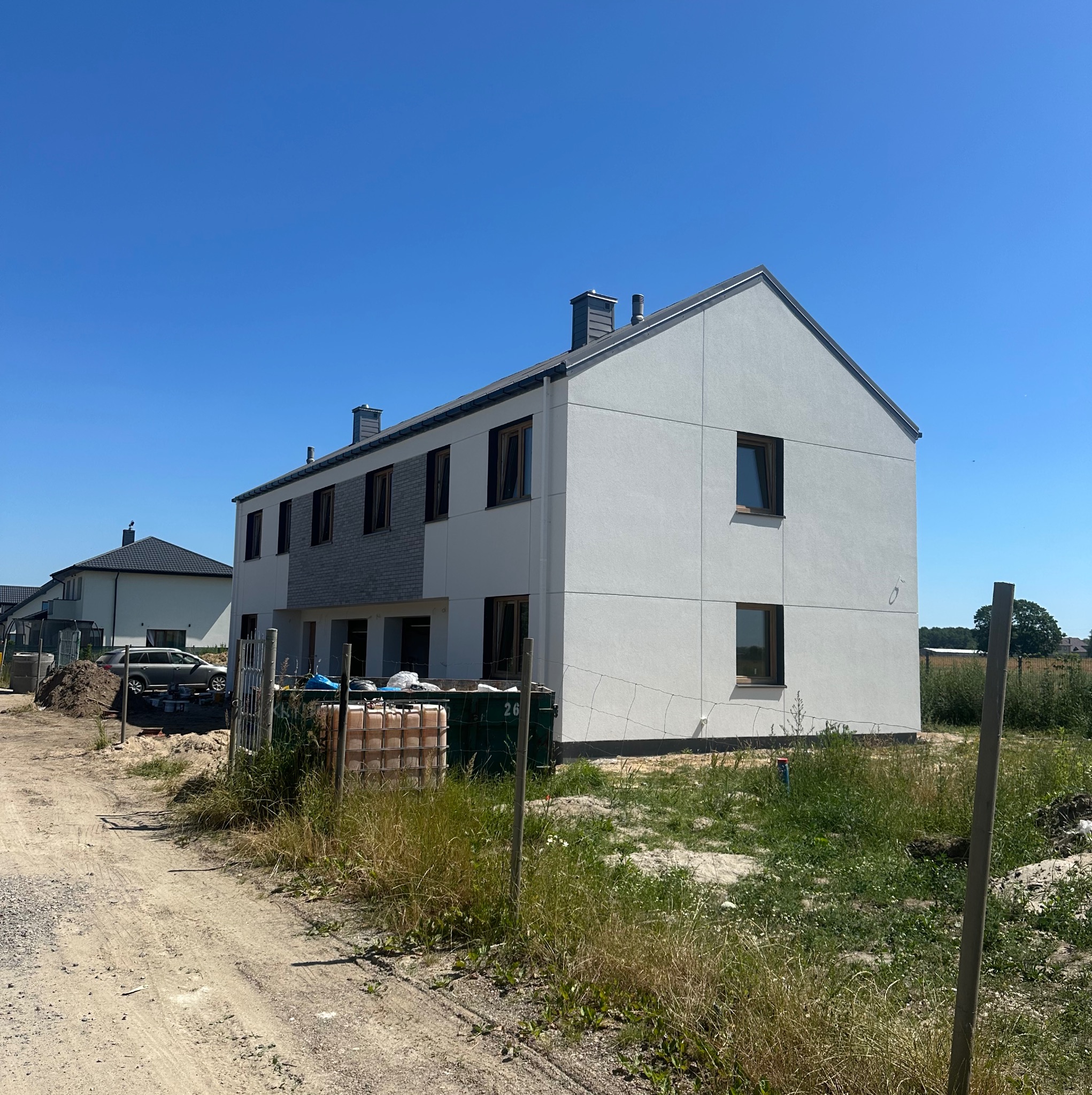 Nowoczesny, dwukondygnacyjny dom z jasną elewacją i szarym fragmentem. Widoczne okna z ciemnymi ramami, kominy i fragmenty otoczenia w słoneczny dzień.