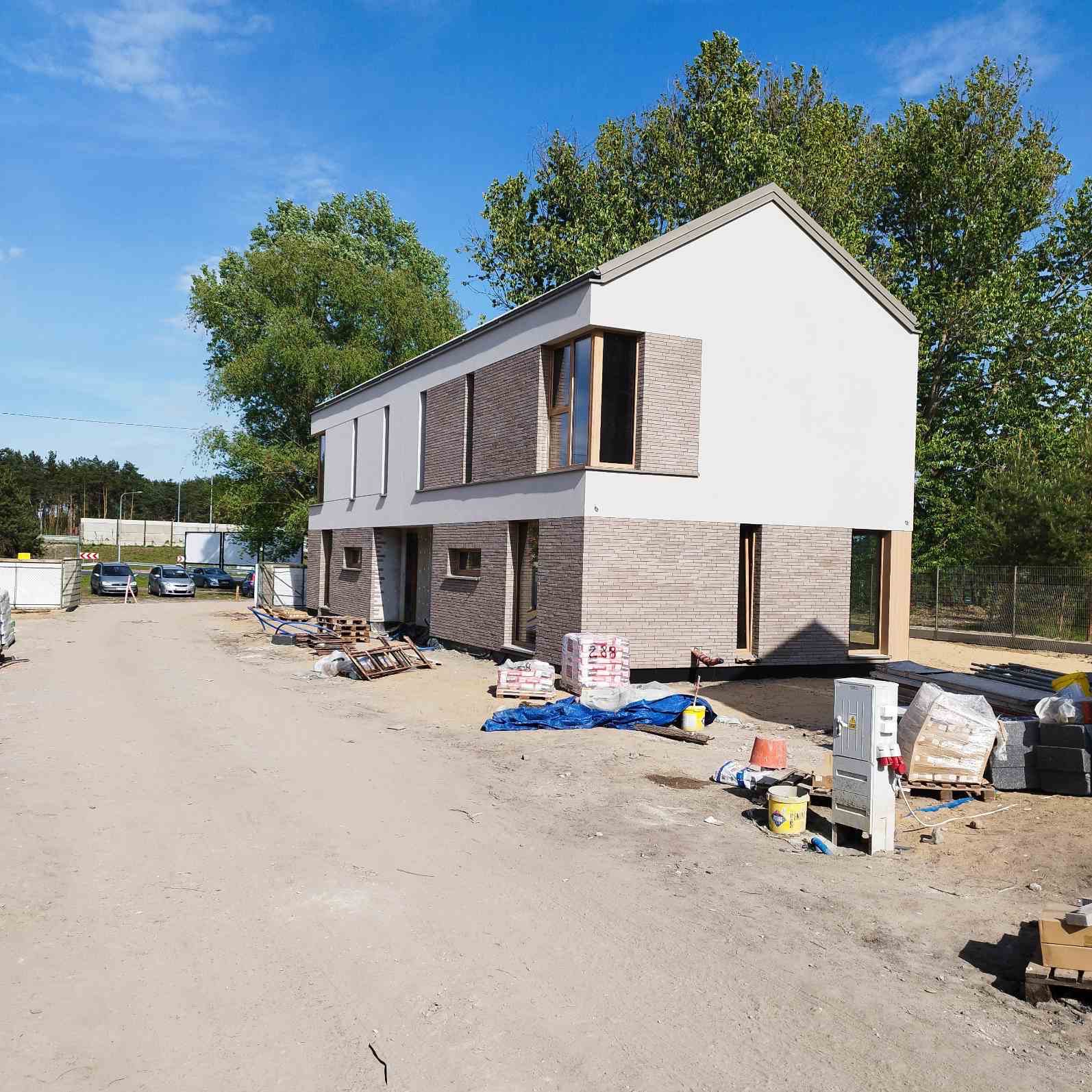 Dwukondygnacyjny dom w trakcie budowy, z częściowo wykończoną elewacją z cegły klinkierowej i białym tynkiem. Widoczne materiały budowlane i tymczasowa skrzynka elektryczna.