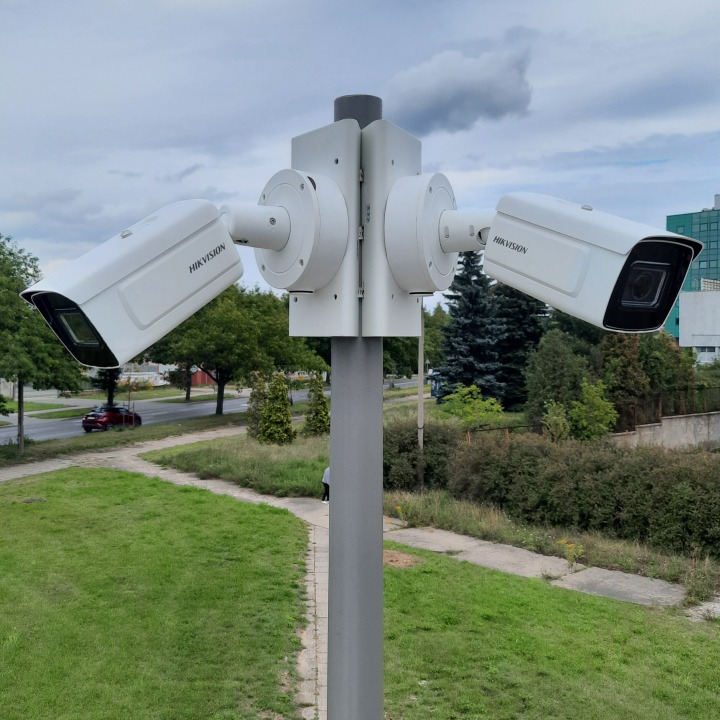 Dwie kamery Hikvision na słupie, skierowane w przeciwne strony, z widokiem na ulicę i zieleń. Monitoring wizyjny w przestrzeni miejskiej.