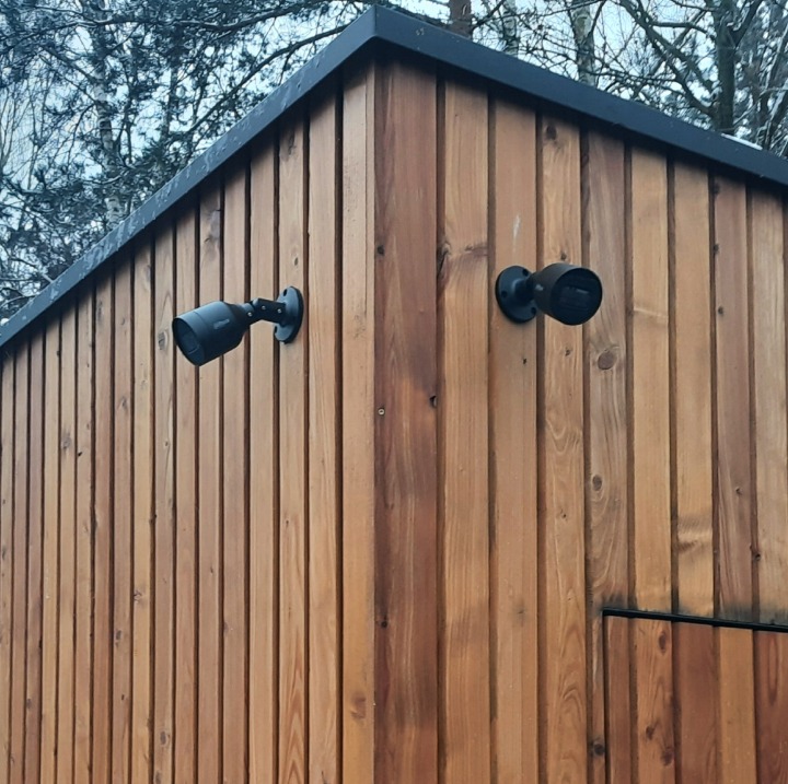 Dwie czarne kamery monitoringu zamontowane na drewnianej elewacji budynku z pionowych desek, widok z dołu, z fragmentem dachu i drzewami w tle.