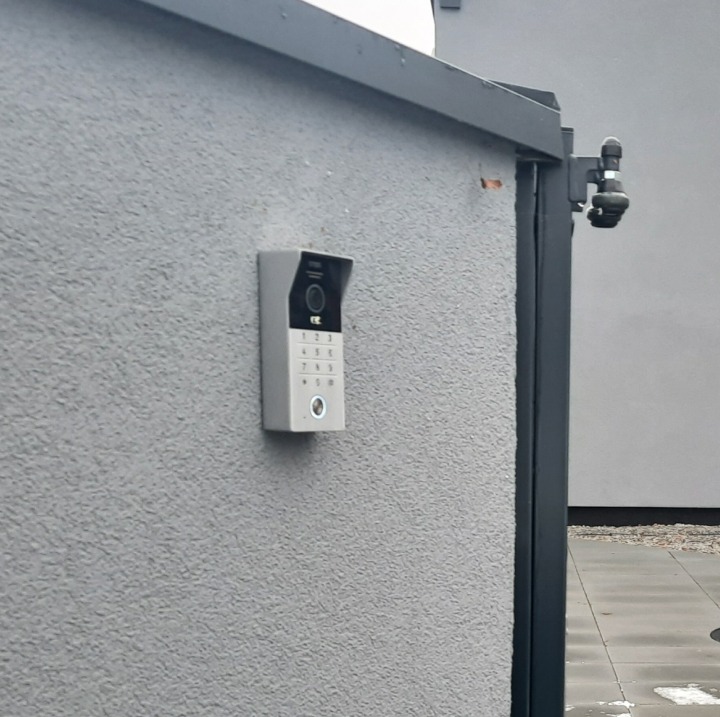 Nowoczesny wideodomofon z klawiaturą numeryczną zamontowany na szarej, teksturowanej ścianie obok czarnej ramy bramy wjazdowej. Widoczna kamera monitoringu.