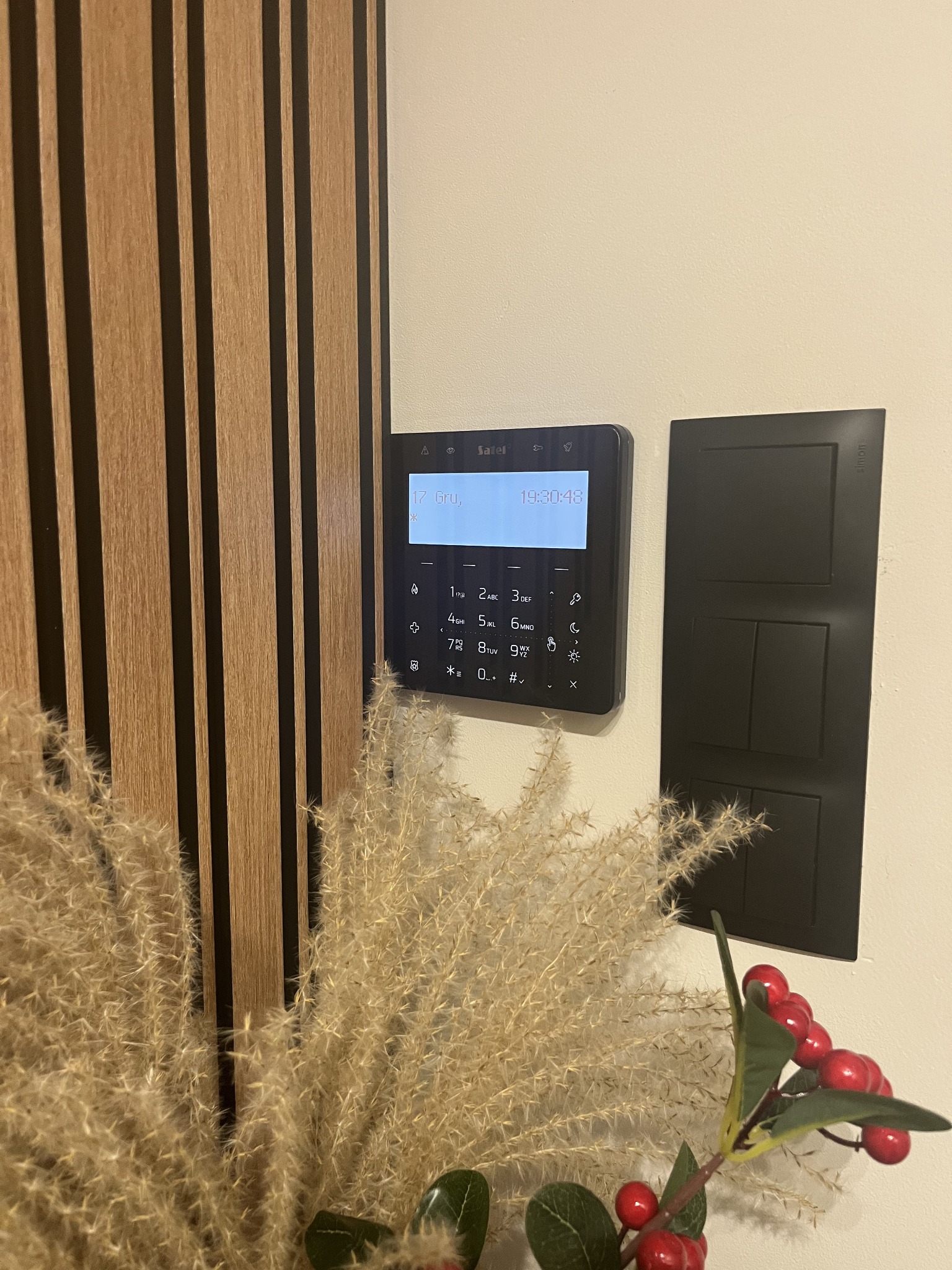 Panel alarmowy Satel z klawiaturą numeryczną, czarny, zamontowany na ścianie obok czarnego panelu z włącznikami światła. Dekoracja z suszonych traw i czerwonych owoców.