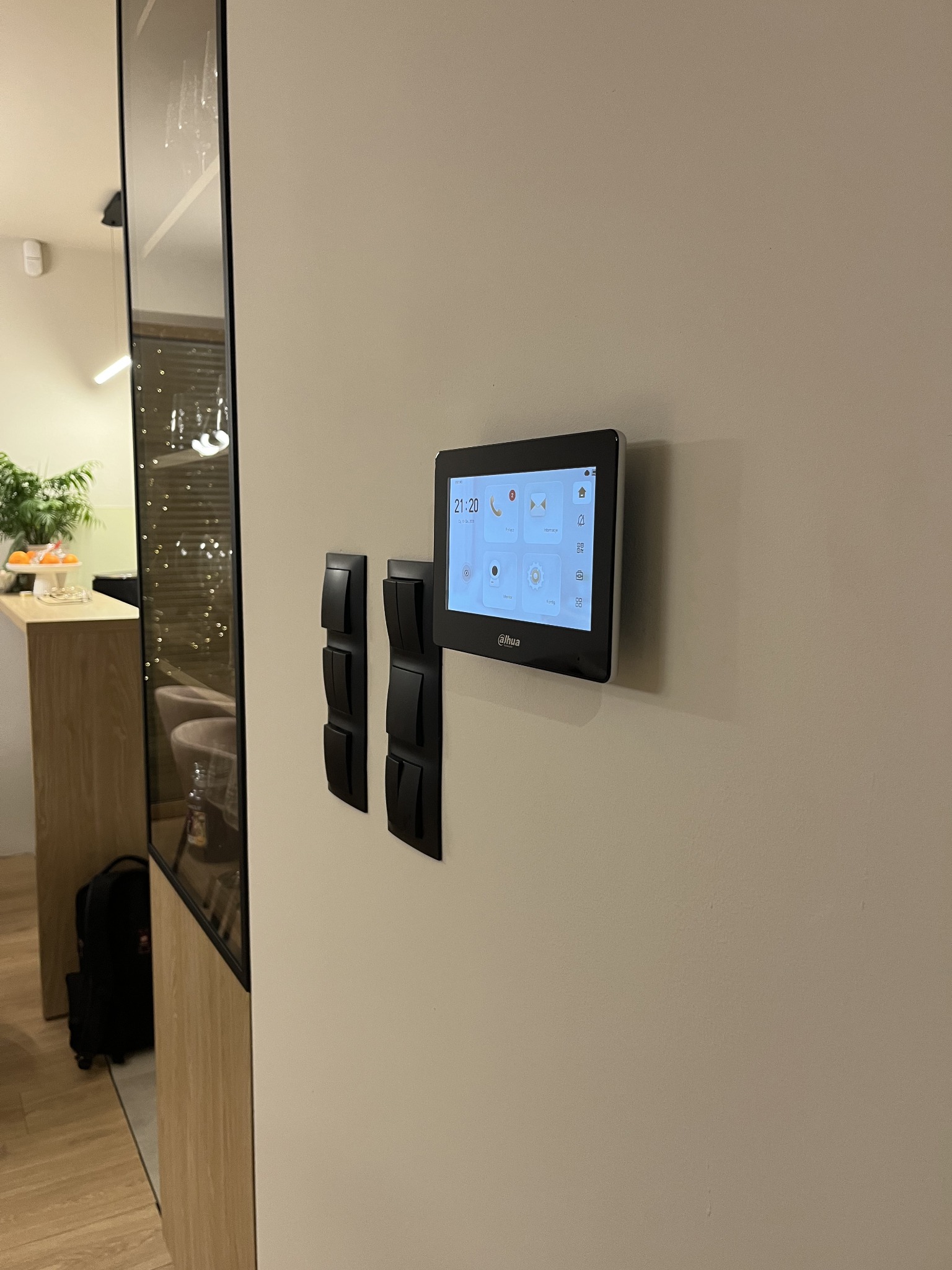 Panel sterowania inteligentnego domu Dahua zamontowany na ścianie obok włączników światła. Widoczny fragment wnętrza z elementami dekoracyjnymi.