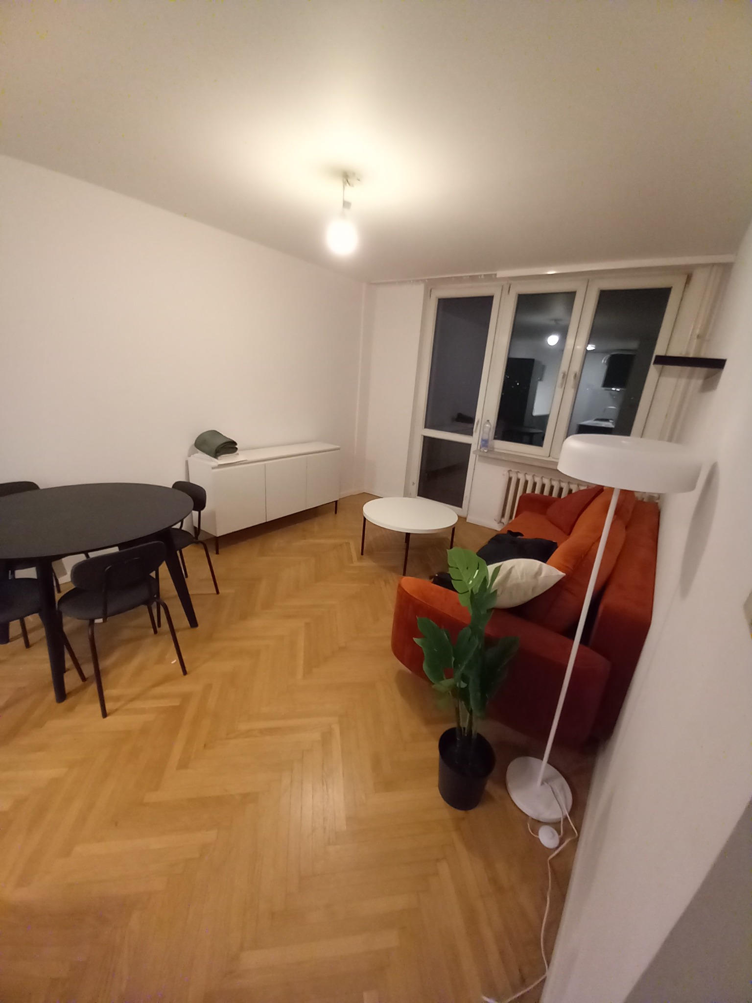 Jasne wnętrze po remoncie w Warszawie: parkiet w jodełkę, pomarańczowa sofa, okrągły stół z krzesłami, biała komoda i lampa podłogowa. Minimalistyczny design.