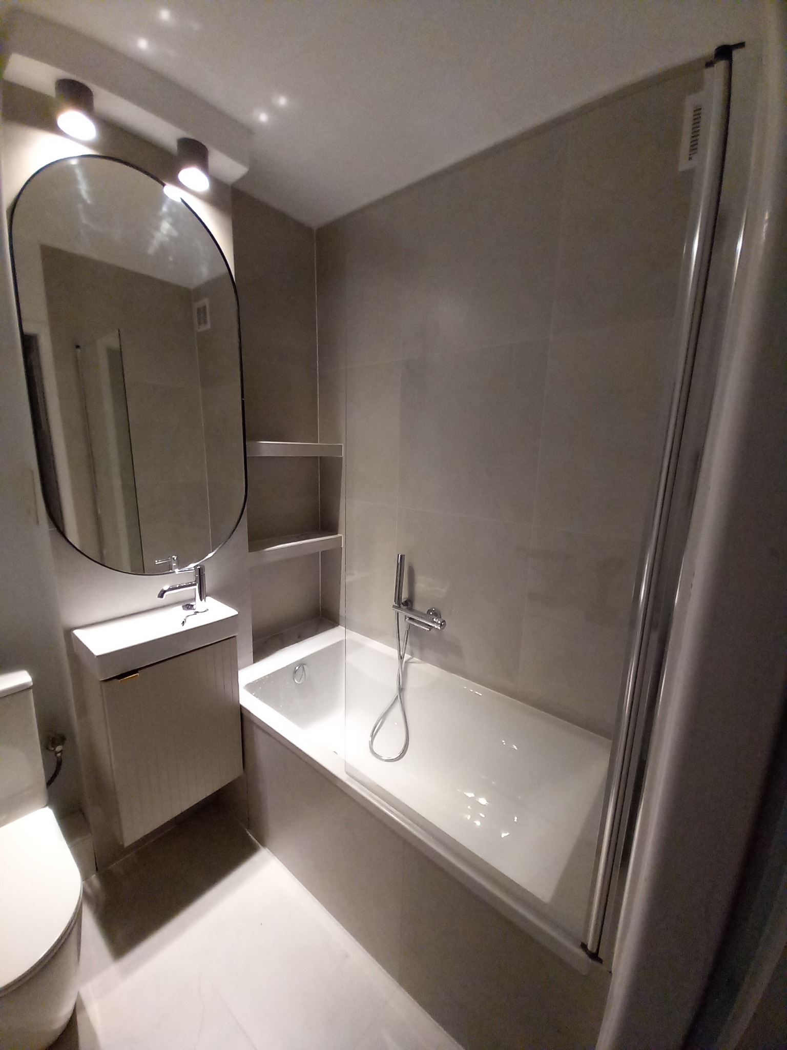 Wykończona łazienka w odcieniach szarości z owalnym lustrem, umywalką z szafką, wanną z parawanem i wnękami na ścianie. Nowoczesne oświetlenie sufitowe.