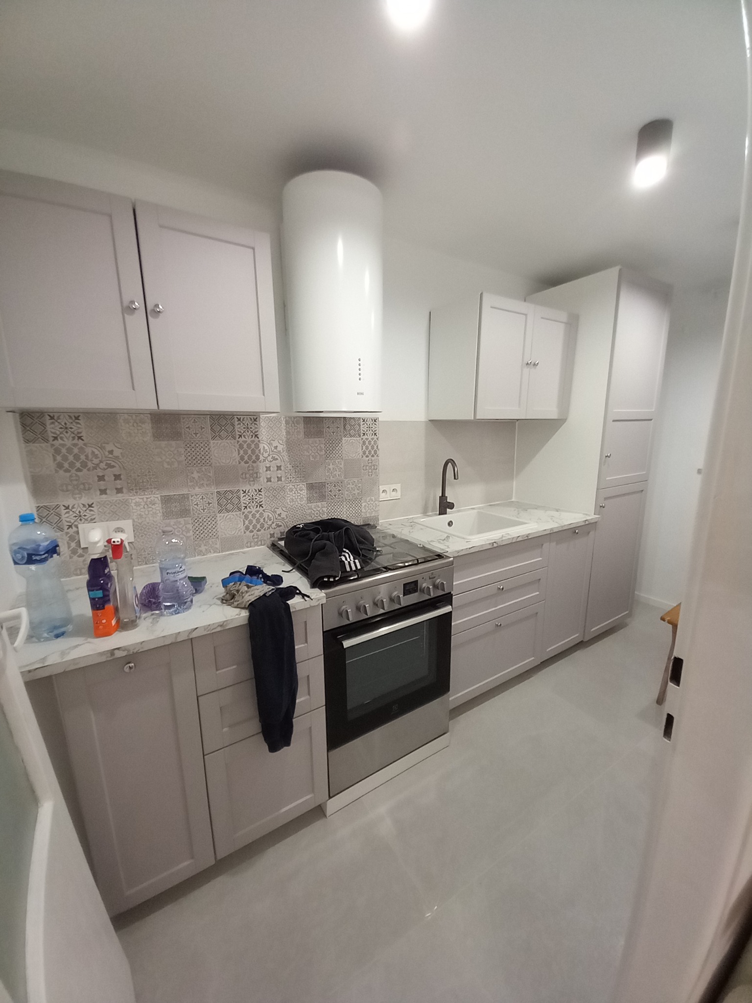 Jasna kuchnia po remoncie: szare szafki, marmurowy blat, wzorzysta ściana nad blatem, biały okap i sprzęt AGD. Widok z perspektywy wejścia, lekki bałagan na blacie.