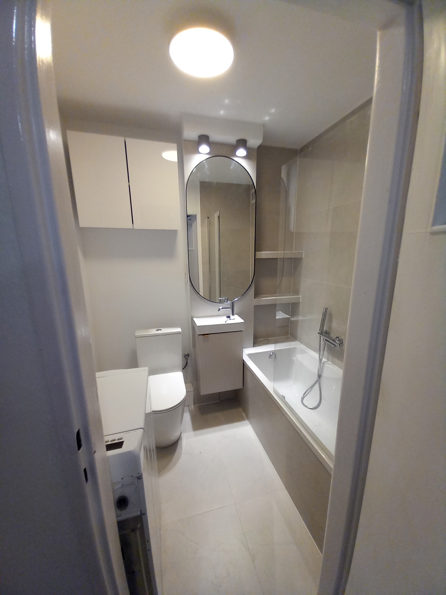 Jasna łazienka po remoncie: biała armatura (toaleta, umywalka, wanna), beżowe płytki, owalne lustro z oświetleniem. Widok zza otwartych drzwi.