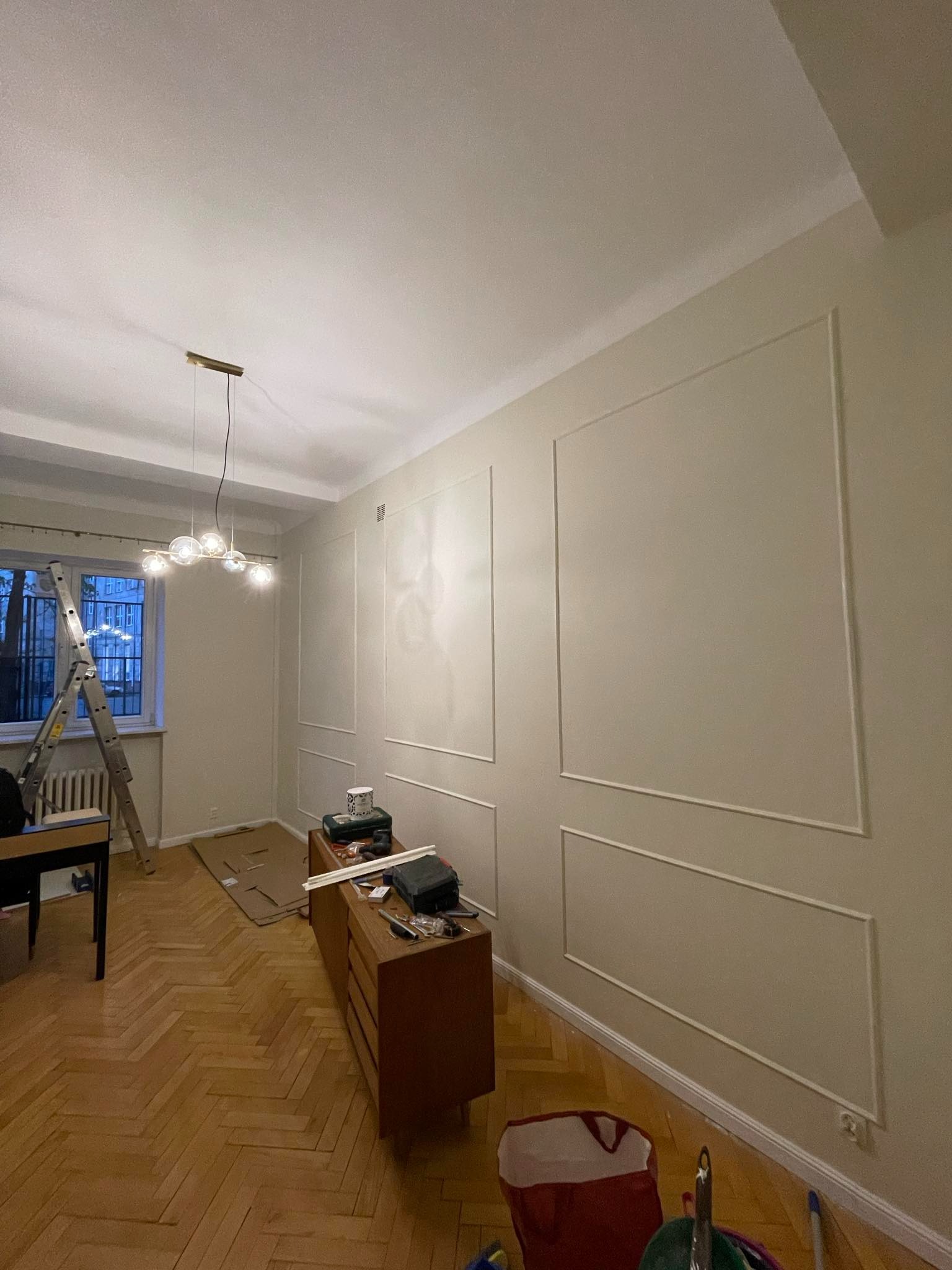 Pokój po dekoracyjnym malowaniu ścian z listwami, drewniana podłoga w jodełkę, drabina przy oknie, komoda z narzędziami. Widoczne ślady prac wykończeniowych.