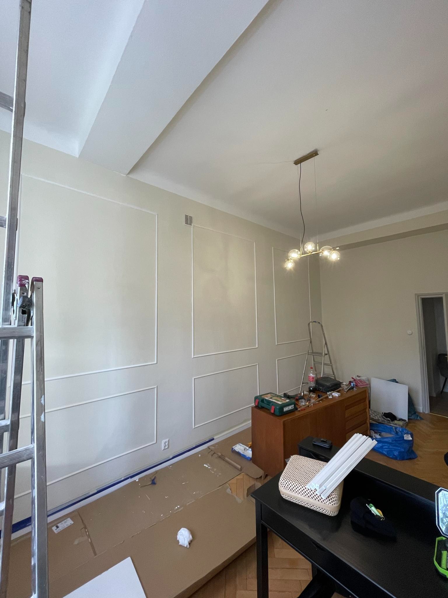 Jasne wnętrze w trakcie dekorowania ścian listwami sztukateryjnymi. Widoczna drabina, narzędzia i zabezpieczona podłoga. Nowoczesna lampa sufitowa.