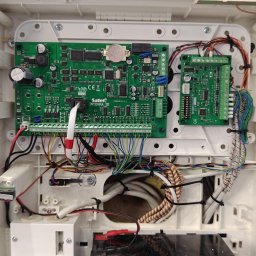 Squdo Technologies Michał Rus - Centrala alarmowa Satel Integra 32 w białej obudowie, widoczne zielone płytki drukowane, liczne przewody i złącza. Detal montażu systemu alarmowego.