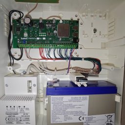 Squdo Technologies Michał Rus - Centrala alarmowa Satel Perfecta 32 LTE w białej obudowie z akumulatorem i transformatorem. Widoczne liczne przewody i podłączenia. Element systemu alarmowego.