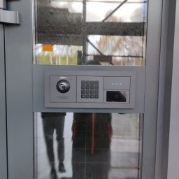 Squdo Technologies Michał Rus - Szary panel domofonu Dahua z kamerą, klawiaturą numeryczną i czytnikiem kart, zamontowany na szklanych drzwiach wejściowych. Widoczne odbicie osoby w szkle.