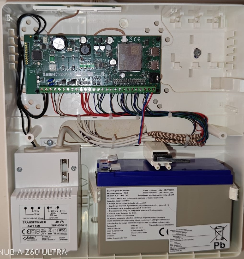 Centrala alarmowa Satel Perfekta z podłączonymi przewodami, akumulatorem i transformatorem w białej obudowie. Widoczne elementy elektroniki i oznaczenia producenta.