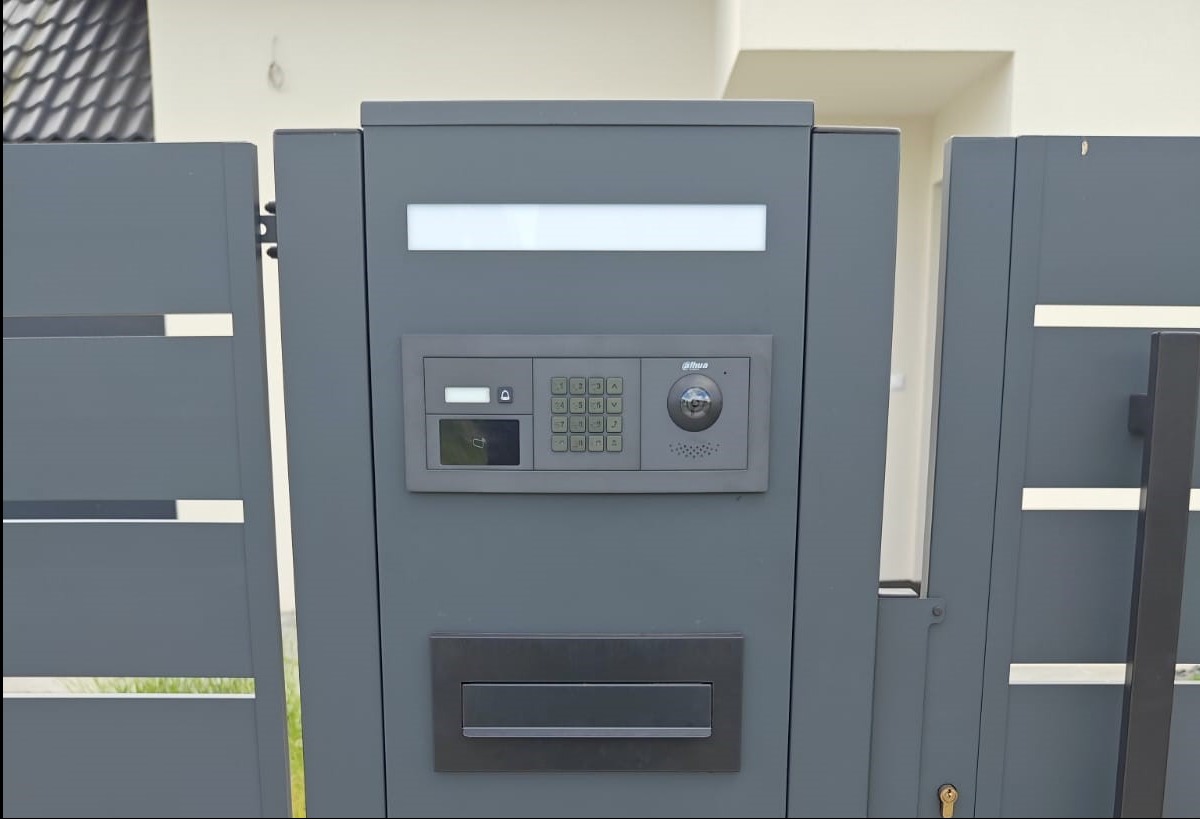 Szara brama z panelem domofonowym Dahua, oświetleniem LED i skrzynką na listy. Nowoczesny design, widok z bliska, lekki cień. Brama częściowo otwarta.