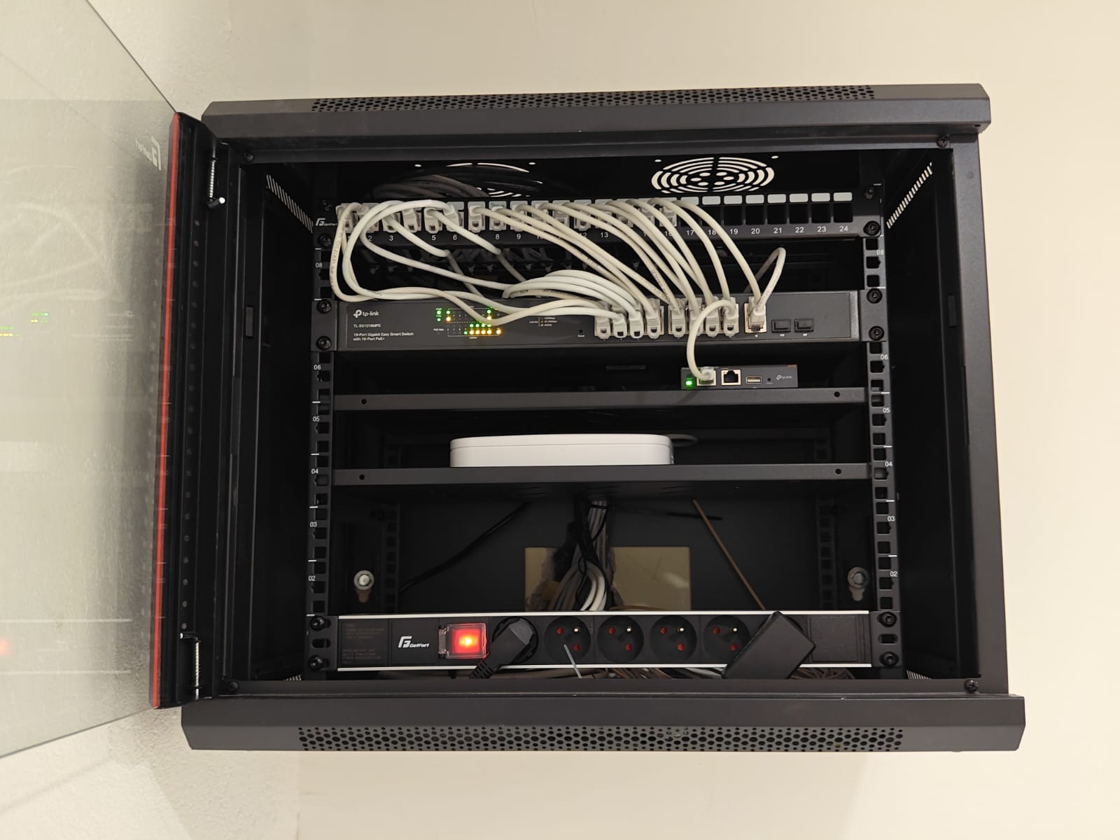 Otwarta szafa rack z zamontowanym routerem TP-Link, switchem, listwą zasilającą i okablowaniem. Widoczne porty Ethernet i diody sygnalizacyjne.