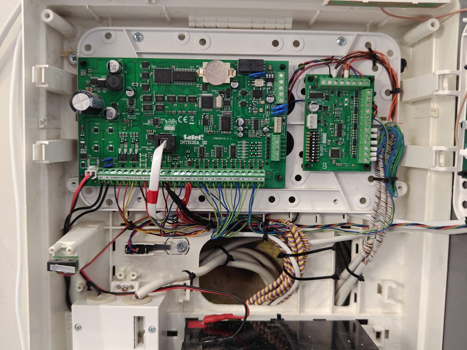 Centrala alarmowa Satel Integra 32 w białej obudowie, widoczne zielone płytki drukowane, liczne przewody i złącza. Detal montażu systemu alarmowego.