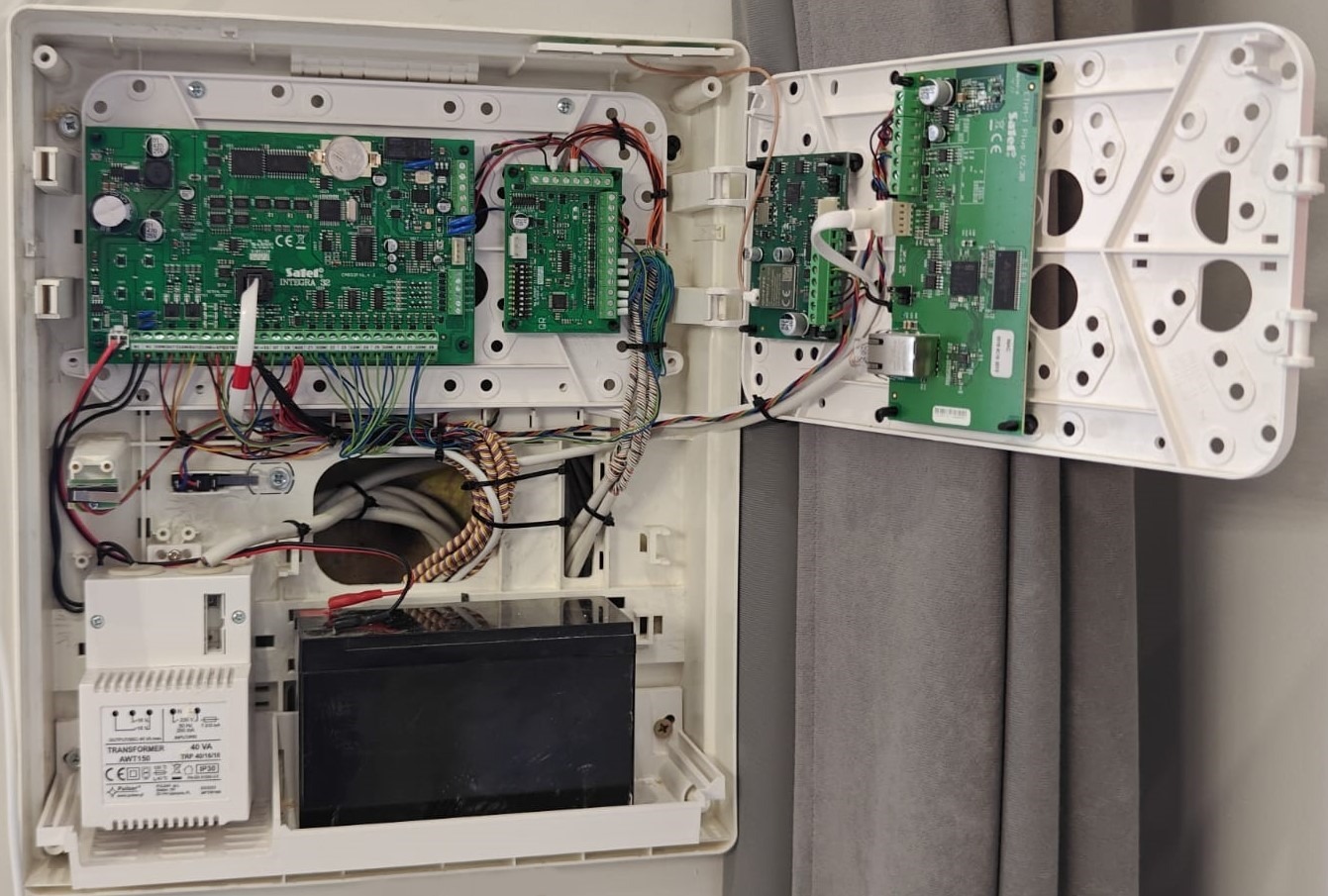 Otwarta obudowa centrali alarmowej z widocznymi płytkami drukowanymi, przewodami i akumulatorem. Instalacja wewnątrz obudowy, widok z bliska.