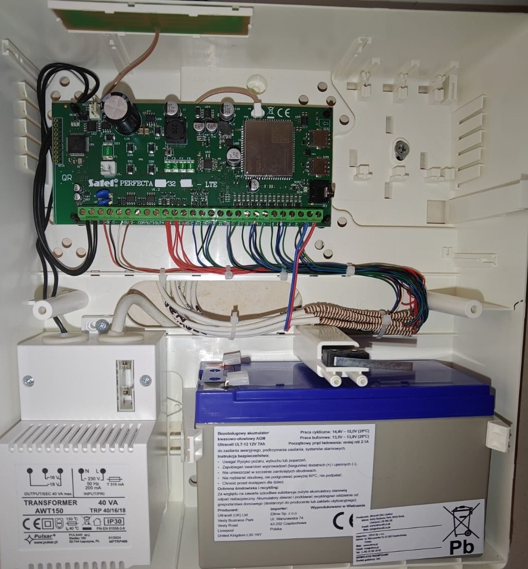 Centrala alarmowa Satel Perfecta 32 LTE w białej obudowie z akumulatorem i transformatorem. Widoczne liczne przewody i podłączenia. Element systemu alarmowego.