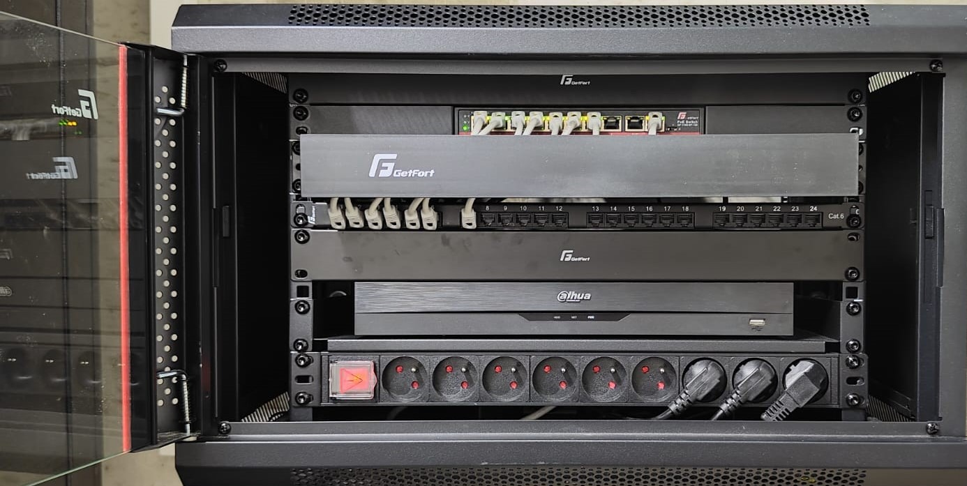 Otwarta szafa rack z zamontowanym switchem, patch panelem, rejestratorem Dahua i listwą zasilającą. Widoczne kable i wtyczki. Sprzęt sieciowy GetFort.