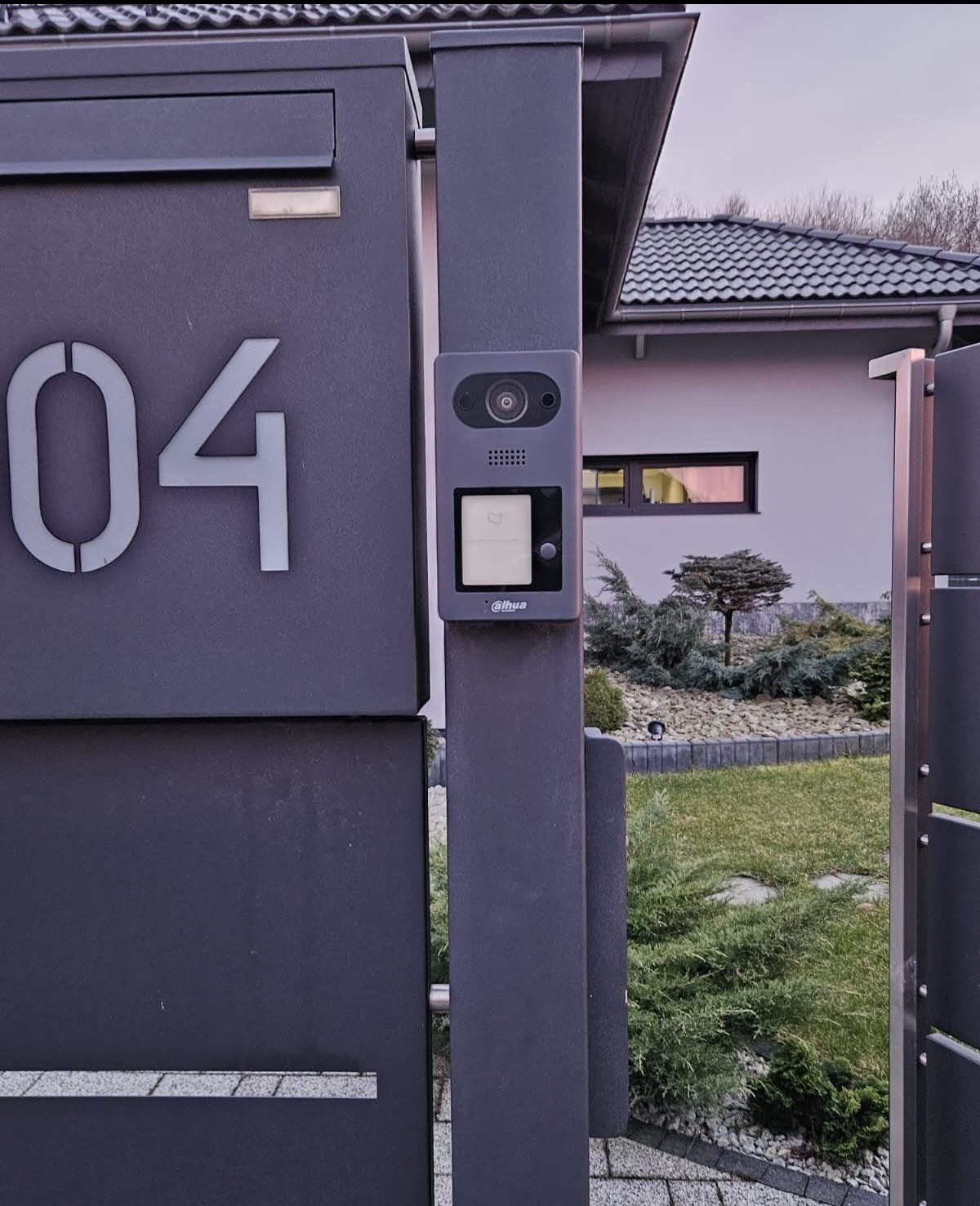 Nowoczesny wideofon Dahua zamontowany na metalowej bramie posesji z numerem 04, na tle domu z ogrodem. Widoczne elementy systemu kontroli dostępu.