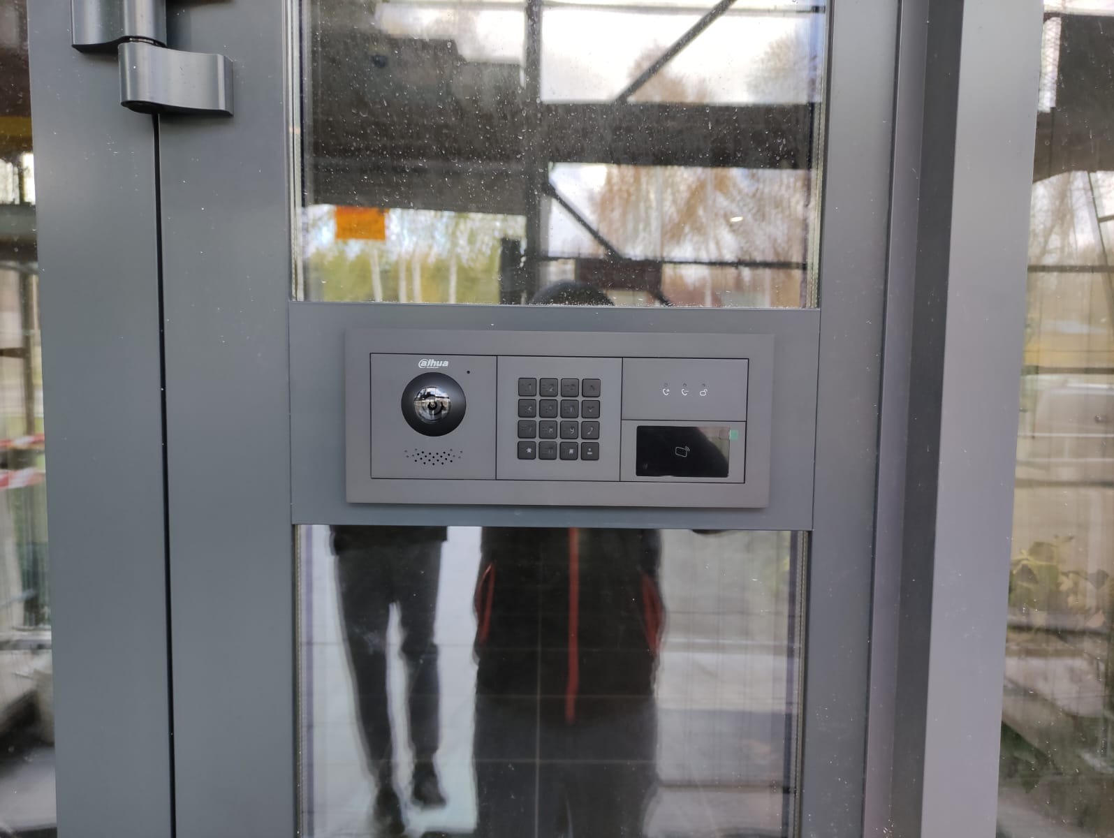 Szary panel domofonu Dahua z kamerą, klawiaturą numeryczną i czytnikiem kart, zamontowany na szklanych drzwiach wejściowych. Widoczne odbicie osoby w szkle.