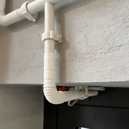 VX Adrian Kurowski - Białe rury przymocowane do szarej ściany, z fragmentem czarnej ramy okiennej w dolnej części kadru. Widoczne uchwyty mocujące rury.