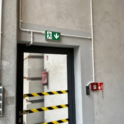 VX Adrian Kurowski - Kluczowy element bezpieczeństwa: znak ewakuacyjny nad drzwiami, czujnik dymu i system kontroli dostępu w nowoczesnym wnętrzu. Widoczny również gaśnica i taśma ostrzegawcza.