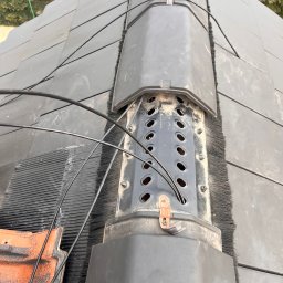 VX Adrian Kurowski - Antena telekomunikacyjna na dachu z czarnej dachówki, z widocznymi kablami i elementami montażowymi. Ujęcie z bliska, podkreślające detale instalacji.