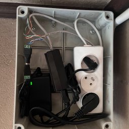 VX Adrian Kurowski - Szara skrzynka instalacyjna z przewodami, zasilaczami i listwą zasilającą. Widoczne kable, wtyczki i złącza. Instalacja elektryczna wewnątrz obudowy.