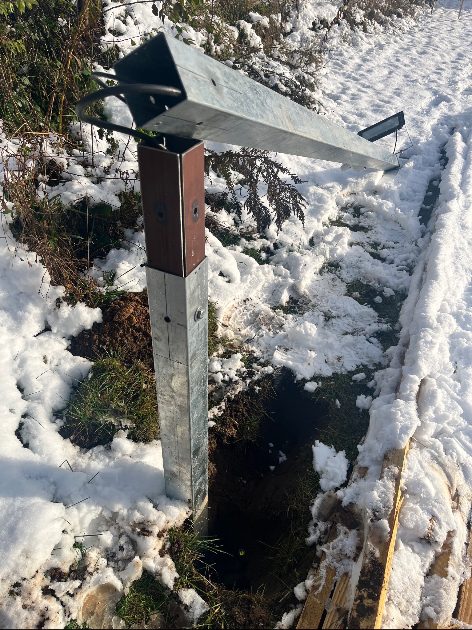 Metalowy słupek z brązowym elementem, prawdopodobnie pod kamerę, wystający z ziemi pokrytej śniegiem. Widoczne ślady prac ziemnych i fragmenty drewnianych desek.