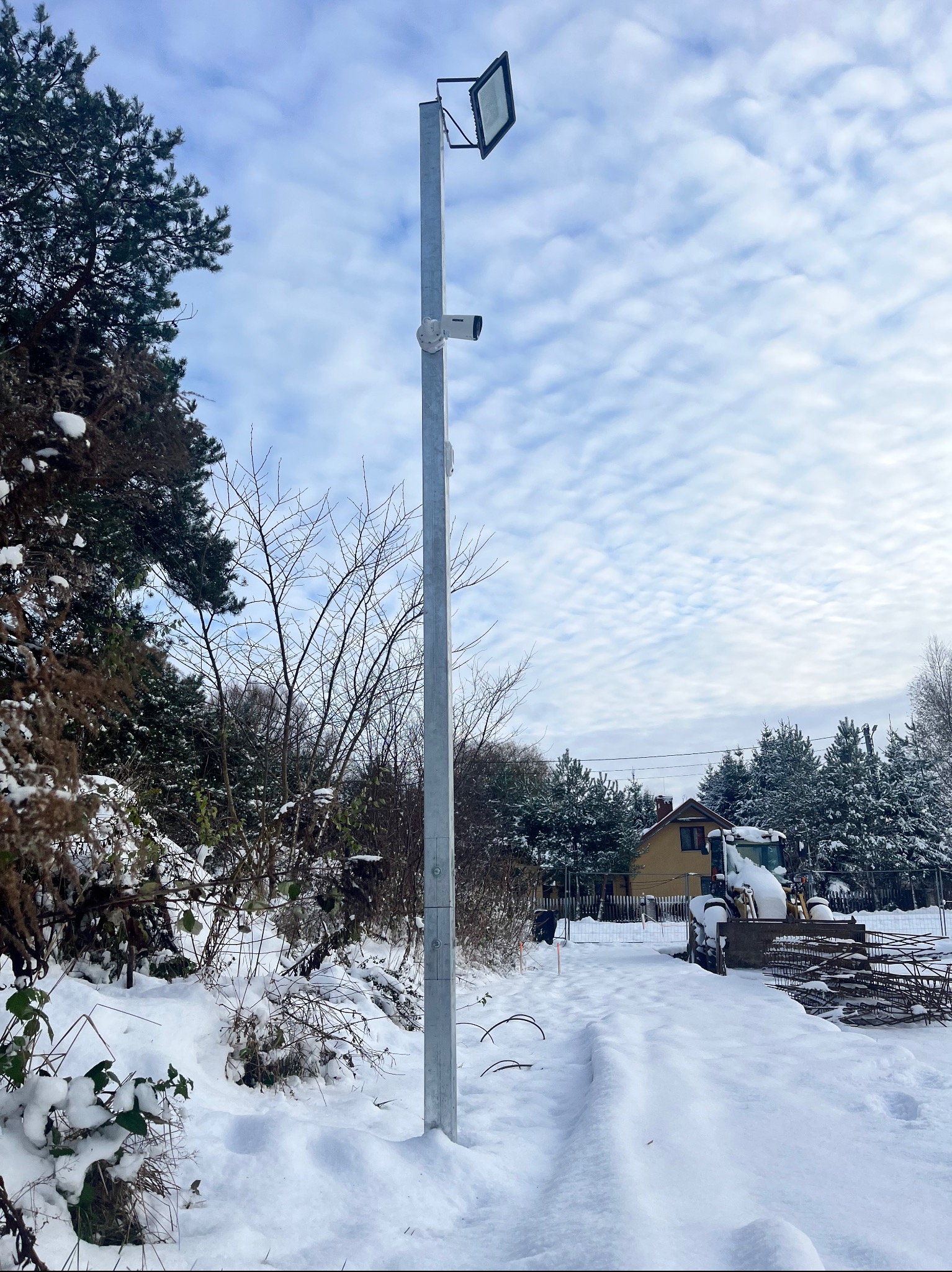 Zimowy krajobraz z metalowym słupem oświetleniowym i kamerą monitoringu, na tle zasypanego śniegiem terenu budowy z widoczną koparką i domem.