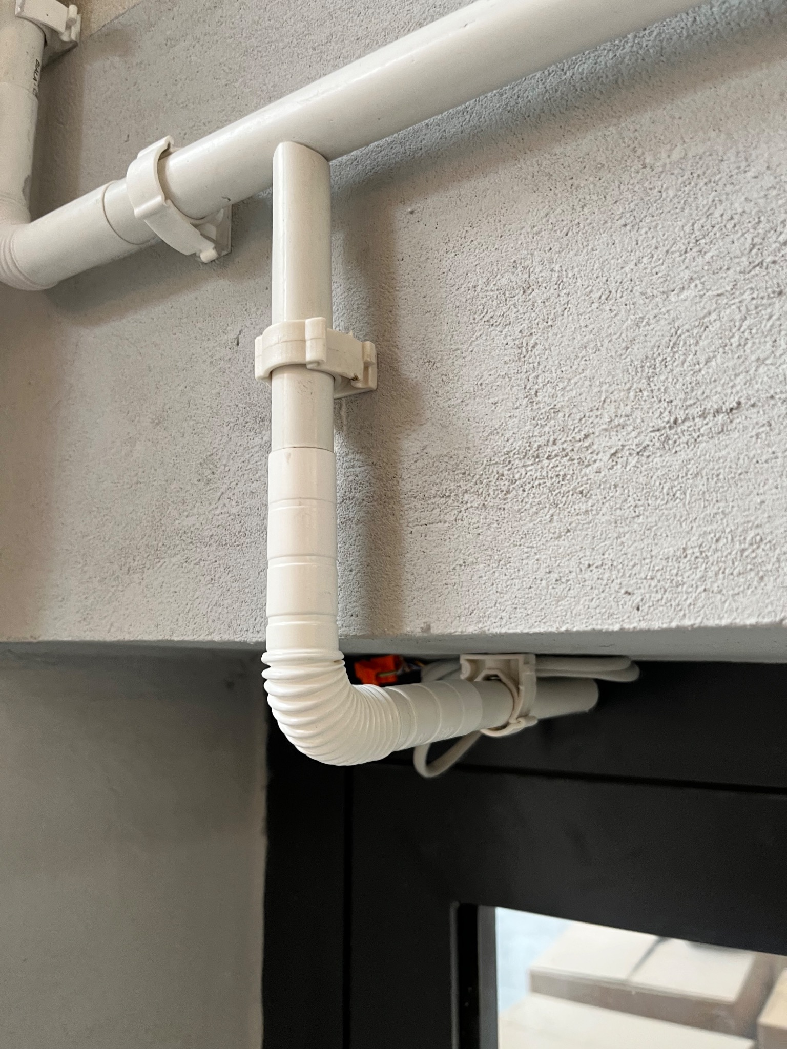 Białe rury przymocowane do szarej ściany, z fragmentem czarnej ramy okiennej w dolnej części kadru. Widoczne uchwyty mocujące rury.