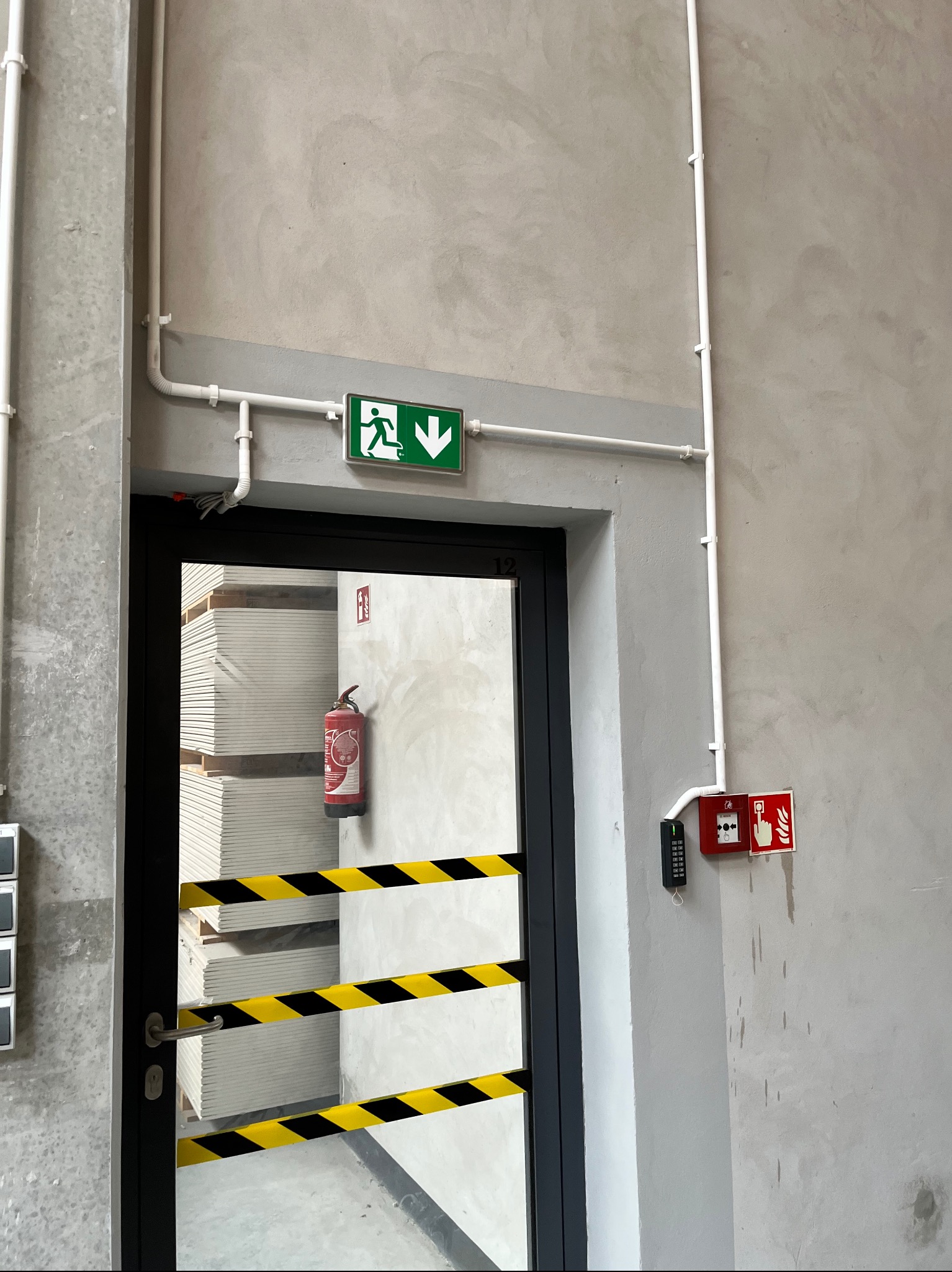 Kluczowy element bezpieczeństwa: znak ewakuacyjny nad drzwiami, czujnik dymu i system kontroli dostępu w nowoczesnym wnętrzu. Widoczny również gaśnica i taśma ostrzegawcza.